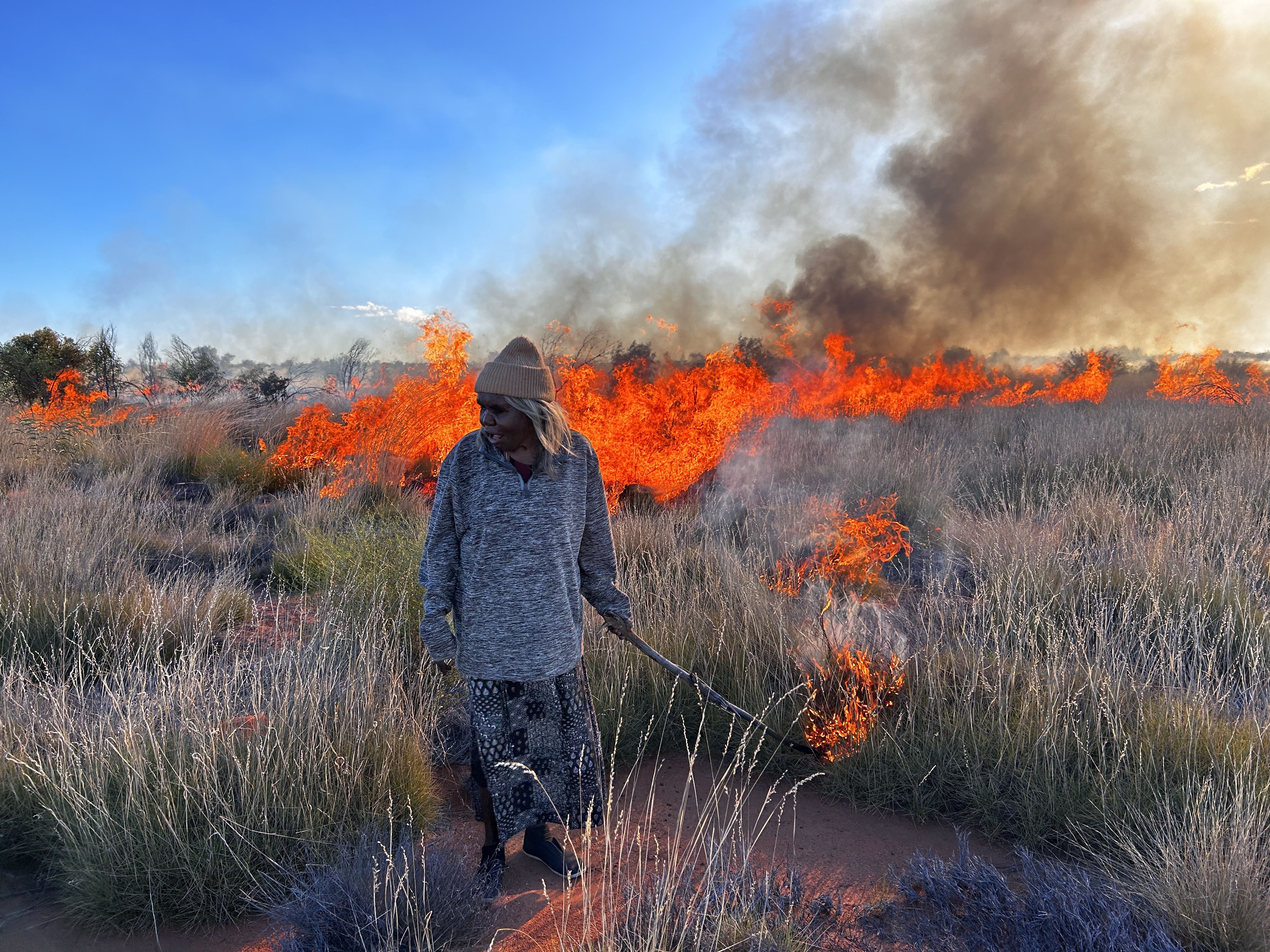 Pintupi Elder Mantua Nangala James holds a firestick and presses it against a clump of dry grass