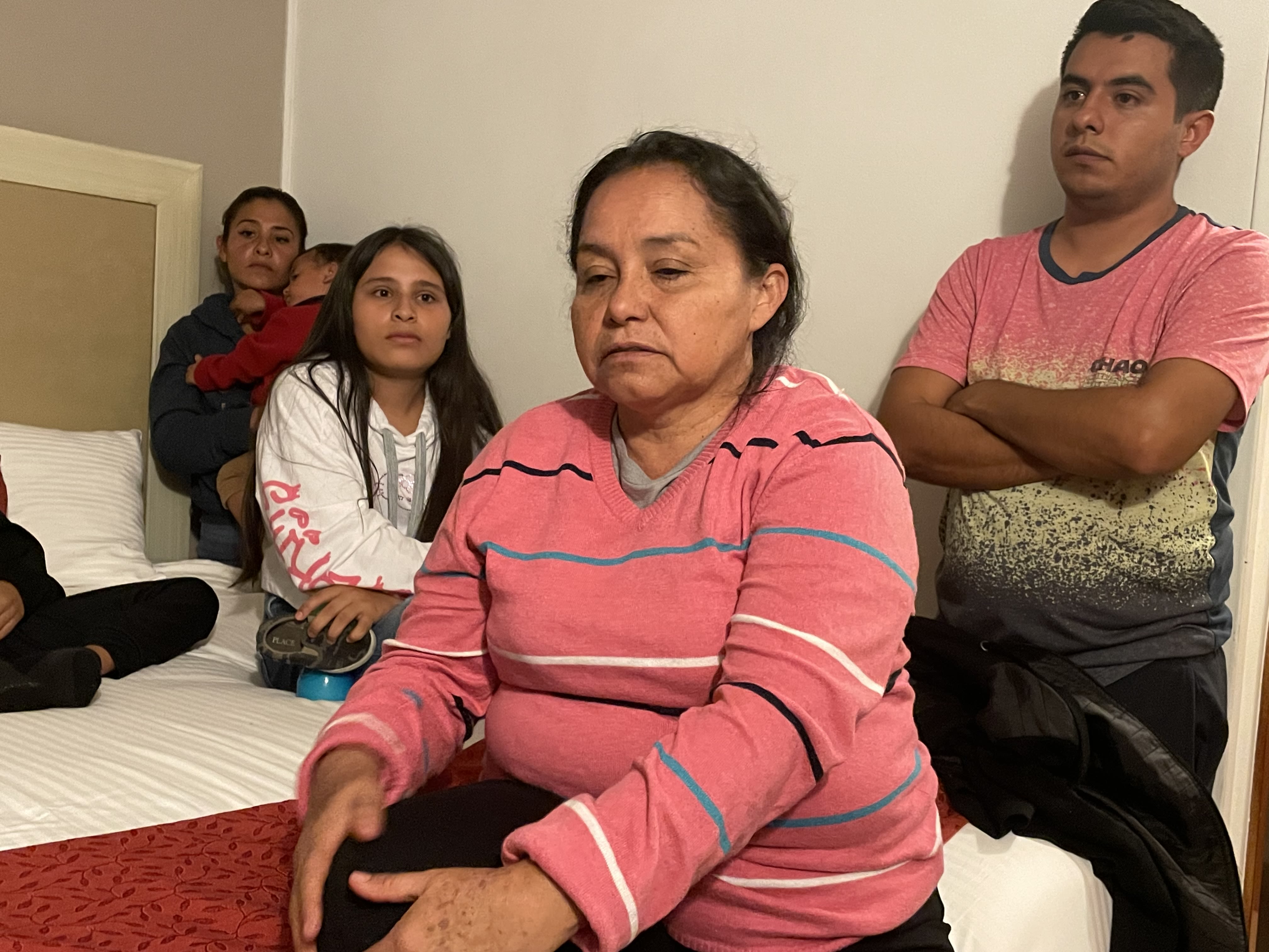 Women sitting on the end of a bed bed. Behind her are a man stands with folded arms, a girl sitting on the bed, and another woman holding a baby